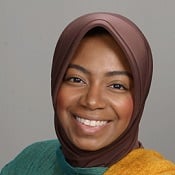 Whitney, a black woman wearing a brown hijab and smiling at the camera. She also wears a green high neck top and a yellow scarf is draped over her right shoulder. 
